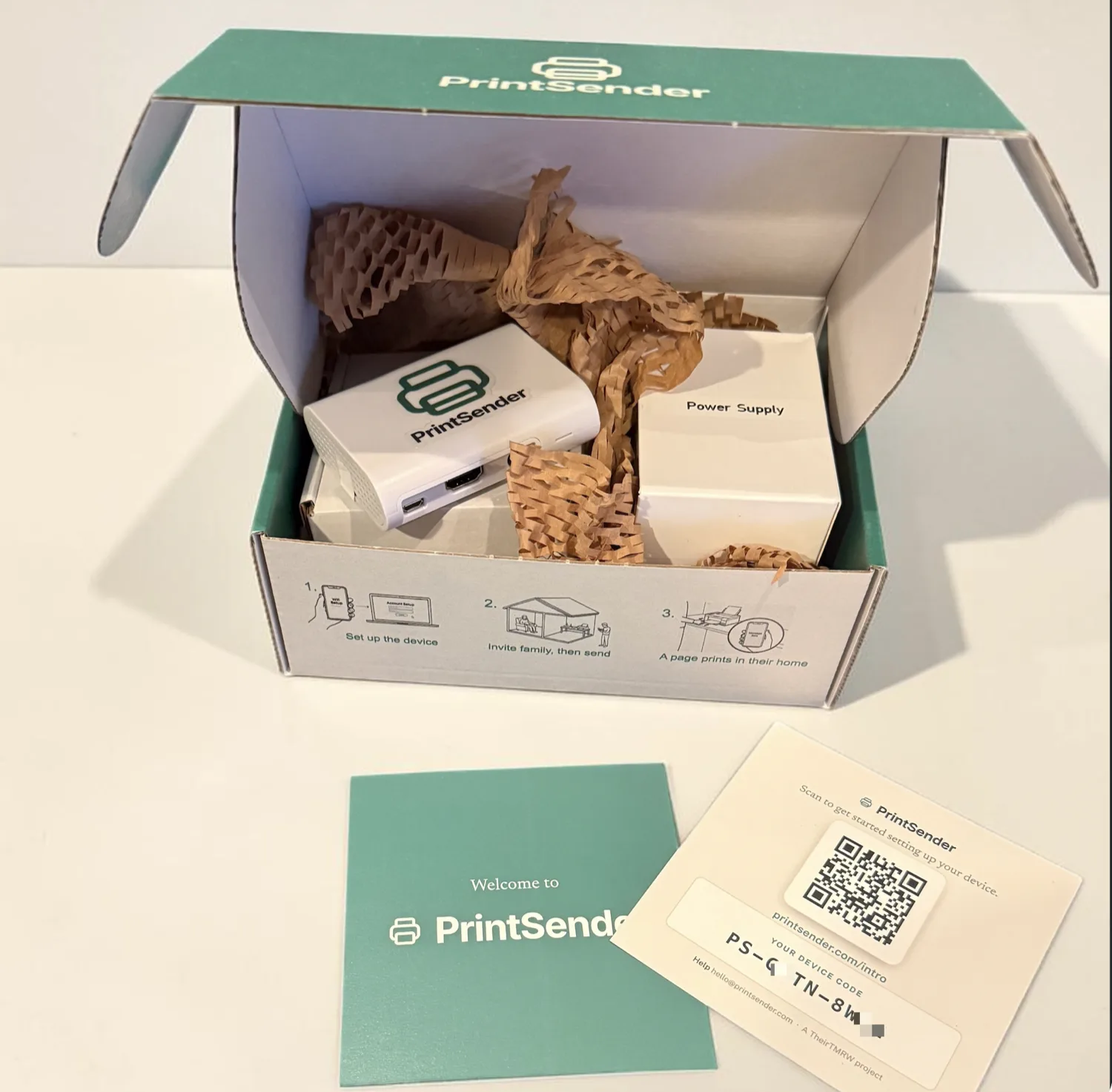 An opened, PrintSender-branded box holding the white device, a Power Supply box, and kraft paper fill, with the three setup steps printed on the box front, plus the green Welcome card and a QR insert card showing the device's setup code
