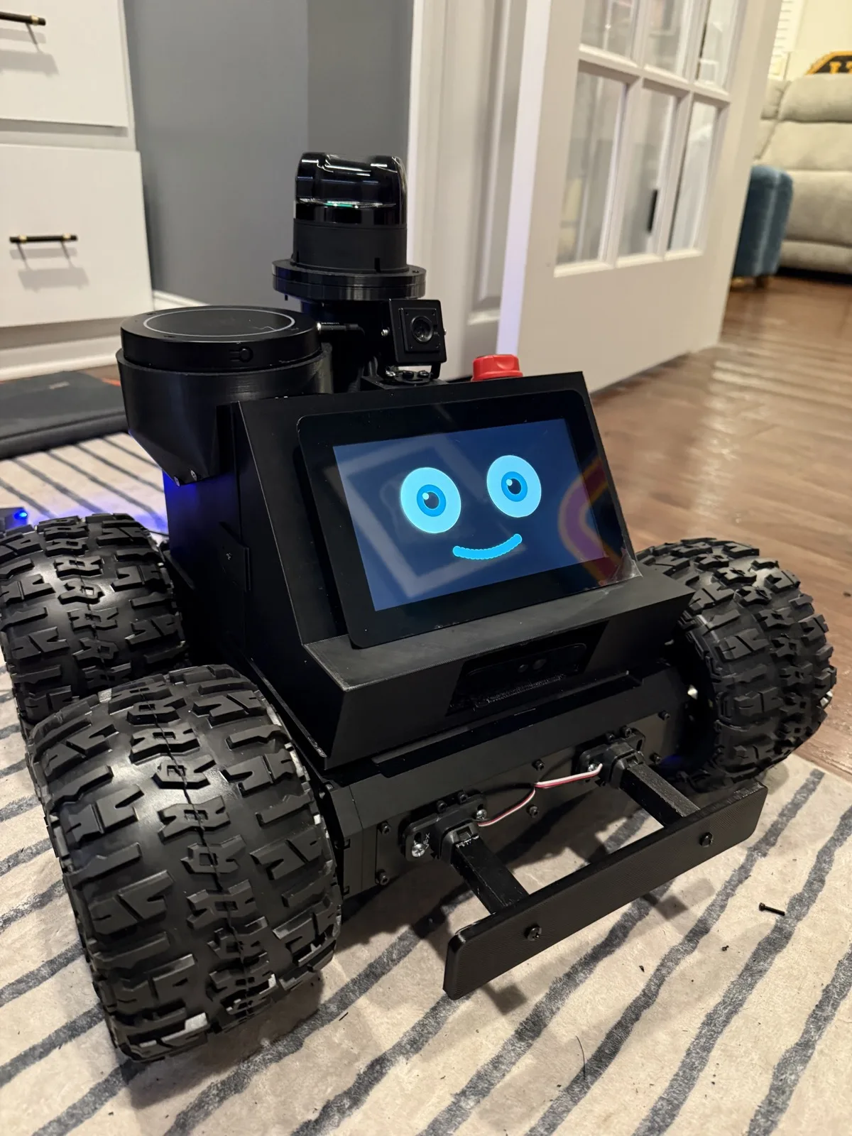 Roger, a black four-wheeled robot with chunky off-road tires, a spinning lidar and a camera on top, a red emergency-stop button, and a 7-inch screen showing a blue smiling face
